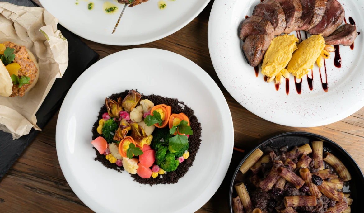 Quattro piatti di cibo gourmet su un tavolo di legno, immortalati durante un servizio fotografico per ristoranti e locali creativi: un piatto di verdure colorate, carne a fette con purea gialla, pasta al forno e un involucro di pasta salata guarnito con erbe.