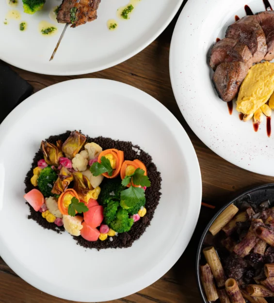 Quattro piatti di cibo gourmet su un tavolo di legno, immortalati durante un servizio fotografico per ristoranti e locali creativi: un piatto di verdure colorate, carne a fette con purea gialla, pasta al forno e un involucro di pasta salata guarnito con erbe.