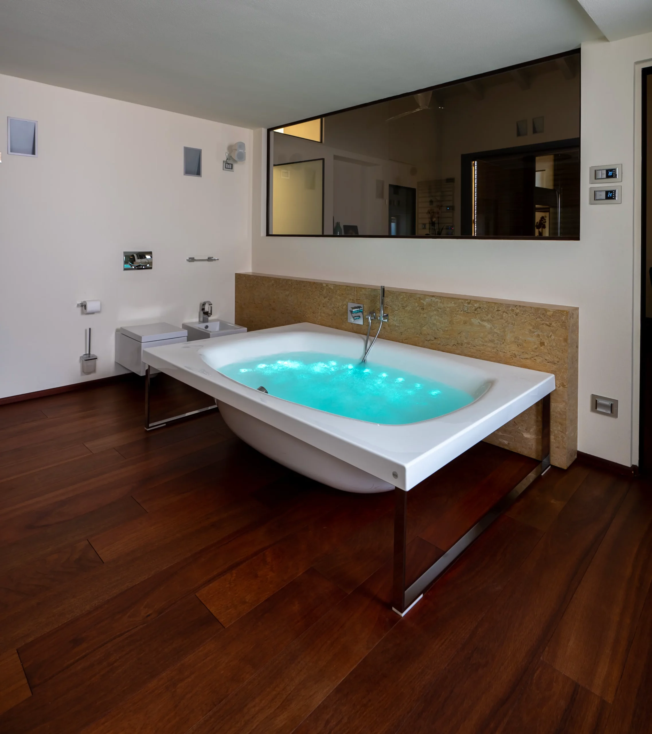 Un bagno moderno con pavimenti in legno scuro, caratterizzato da una vasca bianca con acqua blu brillante, evoca l'eleganza di un bed & breakfast fotografia di lusso Lago di Como. Il bagno è dotato di un grande specchio, di una toilette a muro e di un arredamento minimalista.