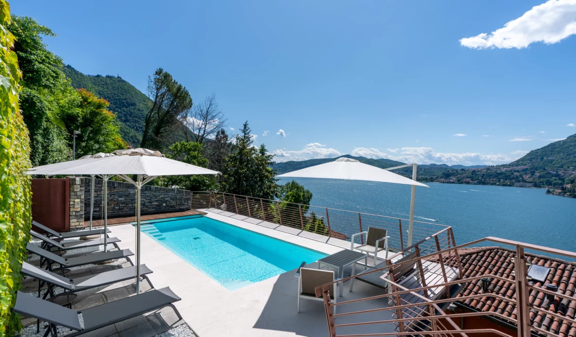 Un moderno ponte della piscina con sedie a sdraio, ombrelloni e posti a sedere si affaccia su un lago panoramico circondato da colline verdi e cielo blu, catturando l'essenza di una fotografia bed & breakfast di lusso sul Lago di Como in un ambiente soleggiato e tranquillo.