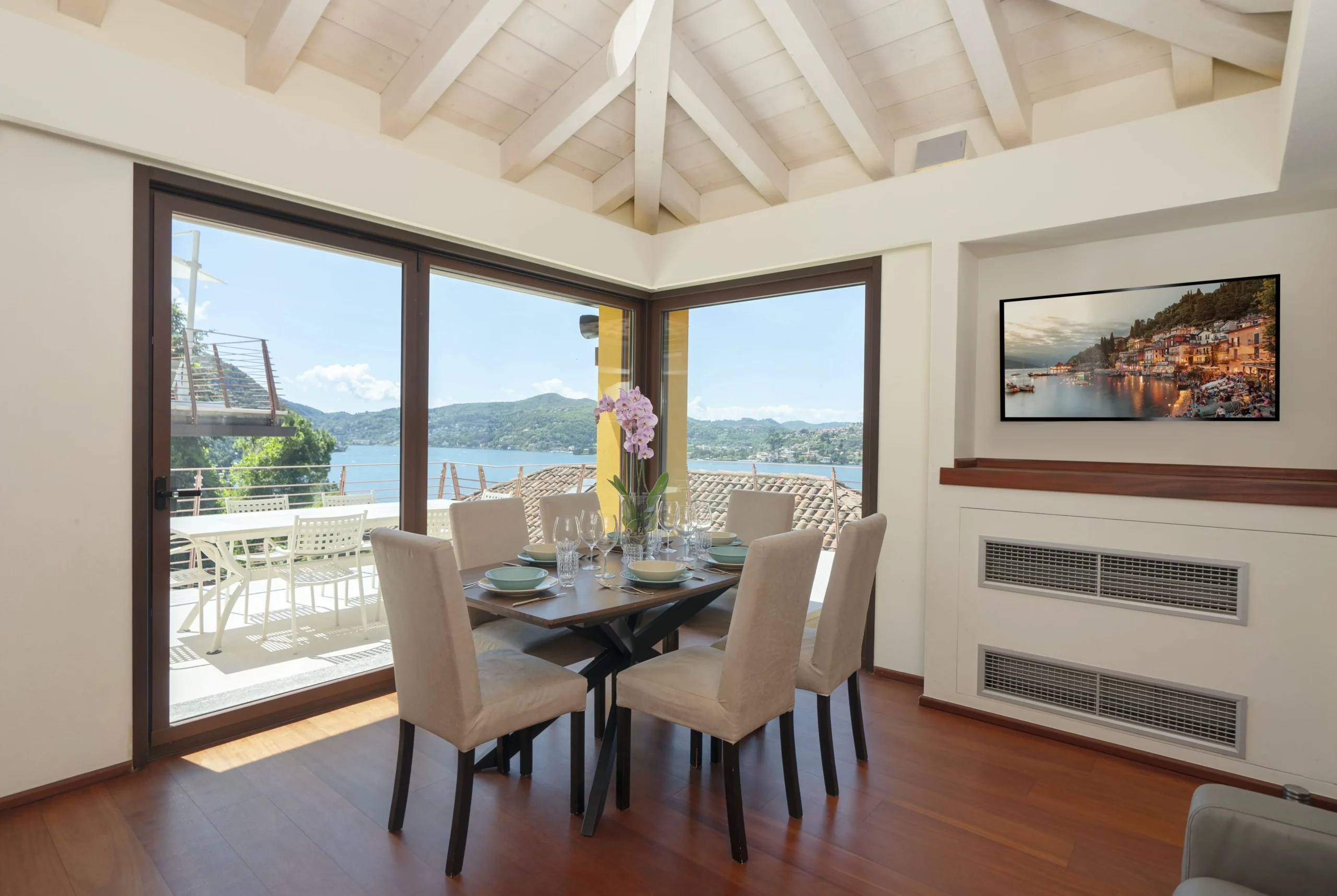 La zona pranzo di un bed & breakfast fotografia di lusso presenta un tavolo in vetro per sei persone, sedie color crema e un centrotavola di orchidee rosa. Grandi finestre incorniciano la vista sul Lago di Como, mentre la TV mostra una scena di un villaggio costiero.
